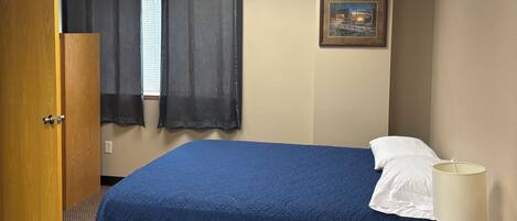 Quarto de hotel com cama, mesa de cabeceira, luminária, porta e janela com cortinas.