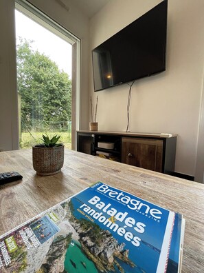 Una habitación con una mesa de madera, una planta en maceta, un control remoto, una revista, un televisor en la pared y vistas a zonas verdes al exterior.