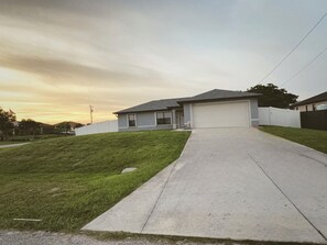 Uma casa com garagem acessível por uma entrada, cercada por um muro branco e um gramado.