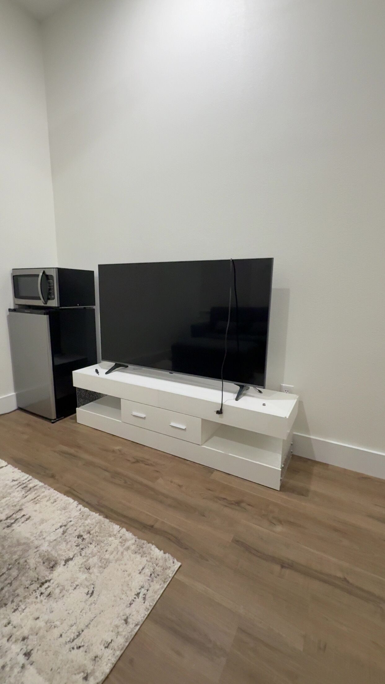 A modern living room with a flat-screen TV on a white stand, a microwave above it, and a small refrigerator to the left.