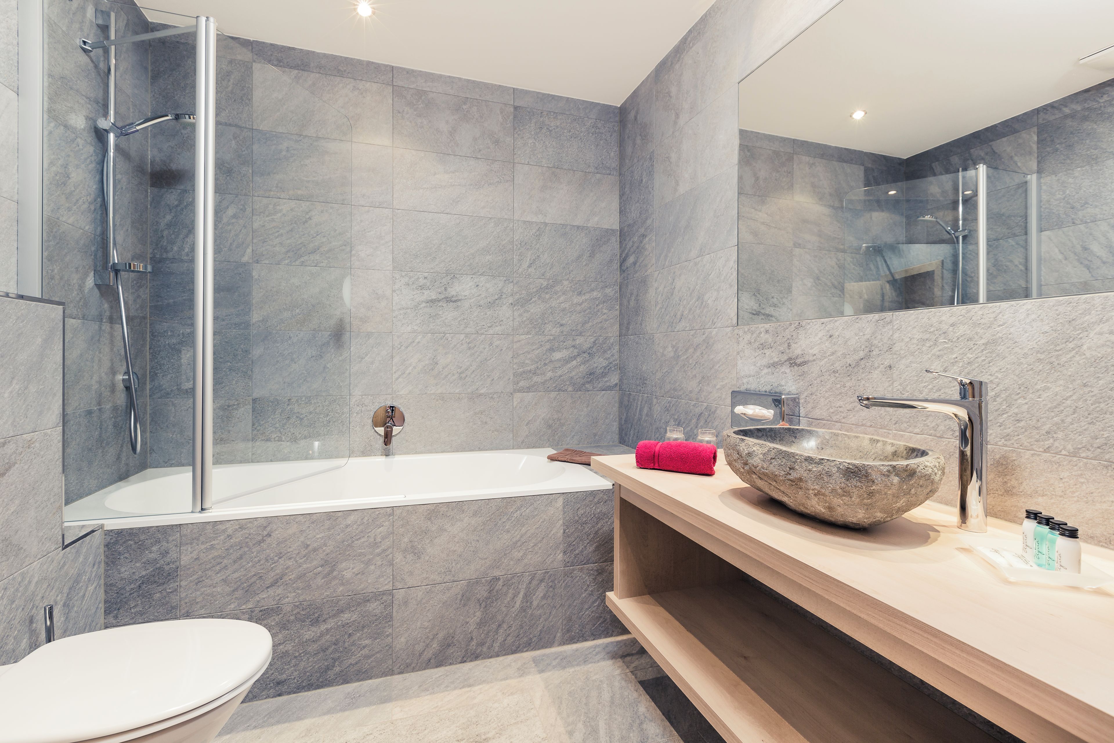 Un baño moderno con ducha envuelta en vidrio, una bañera blanca y un espejo grande sobre un tocador de madera con lavamanos de piedra.