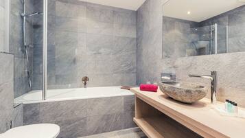 Un baño moderno con ducha envuelta en vidrio, una bañera blanca y un espejo grande sobre un tocador de madera con lavamanos de piedra.