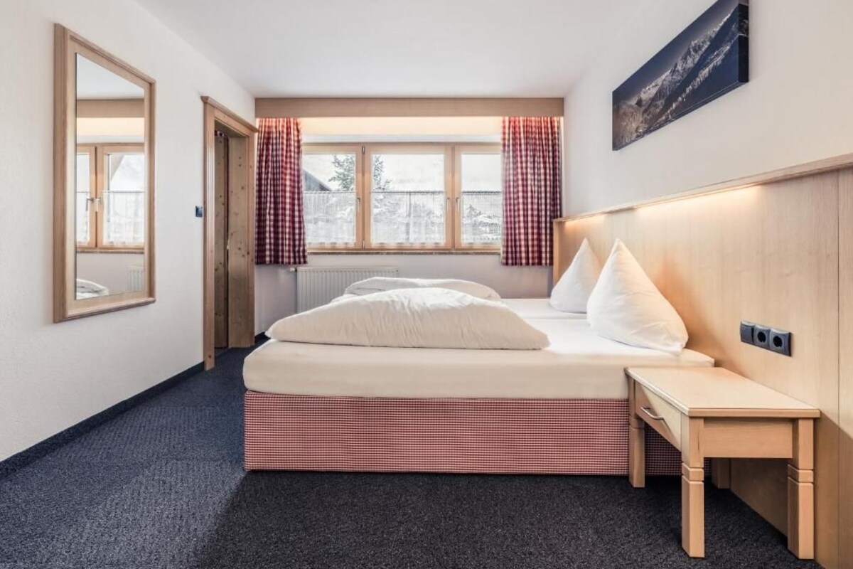 Een hotelkamer met een bed, nachtkastje, raam met gordijnen, radiator en een aan de muur hangend schilderij.