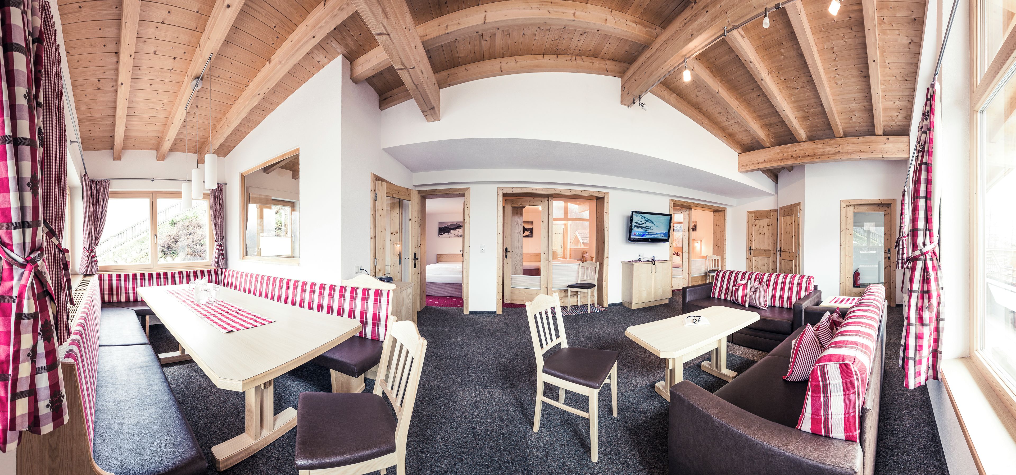 Una habitación amplia con techo de madera alto, un ventanal, televisor y una zona de estar con sofá y sillones.