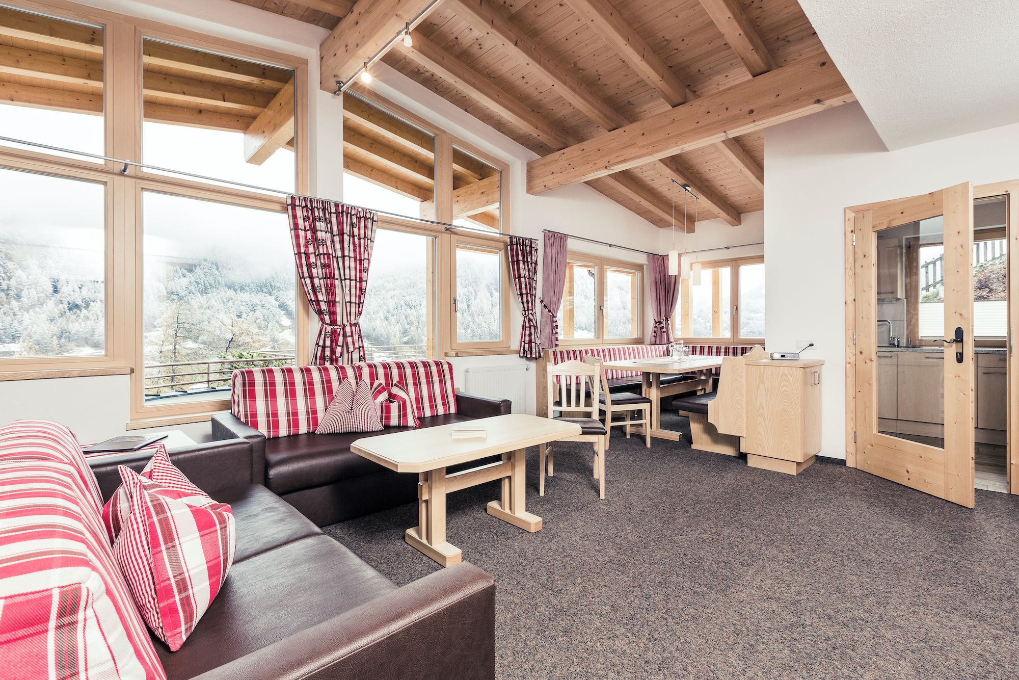 Ein gemütliches Zimmer mit einer Couch, einem Tisch und Stühlen, großen Fenstern mit Vorhängen und Blick auf eine verschneite Landschaft.