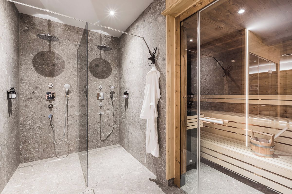 Un baño moderno con pared de piedra, cabina de ducha de vidrio y sauna de madera visible a través del cristal.