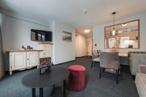 A modern living room with a flat-screen TV mounted on the wall, a round table with a red ottoman, and a seating area with a sofa and chairs.