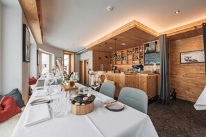 Un restaurante con una larga mesa de comedor dispuesta para una comida, detalles de madera y una zona de bar con estantes llenos de botellas.