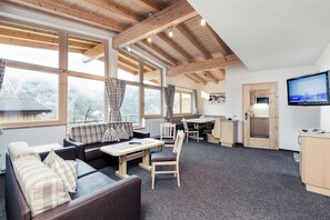 A modern interior with a sofa, dining table, and kitchen area, featuring large windows and wooden beams. - Leni Mountain Apartments (Soelden)