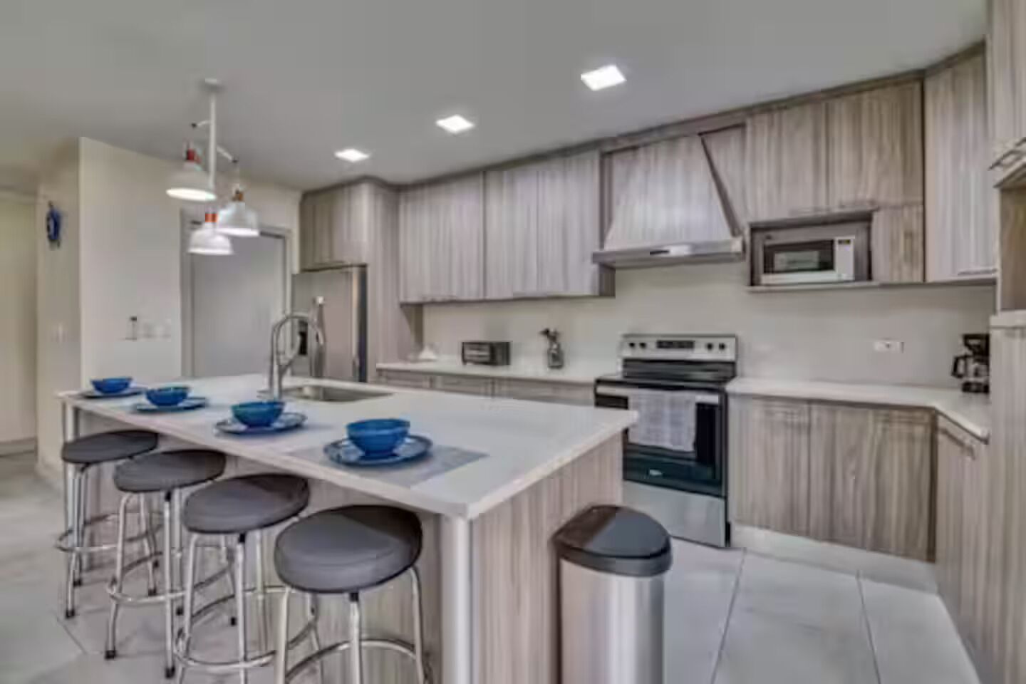 Una cocina moderna con una isla central, taburetes de bar, electrodomésticos de acero inoxidable y armarios de madera clara.