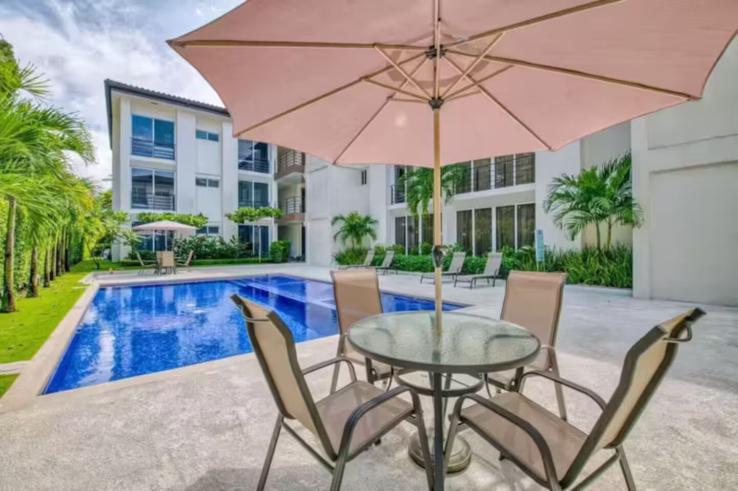 A swimming pool with lounge chairs and an umbrella, surrounded by greenery and modern buildings.