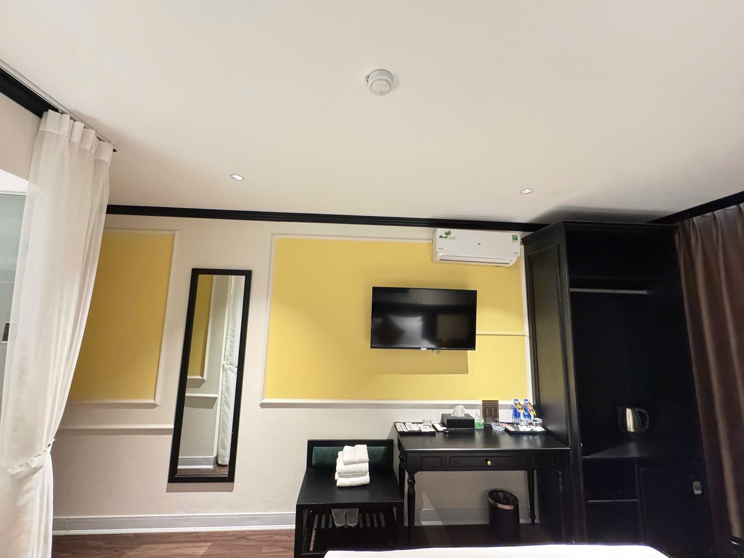 A hotel room with a bed, a desk with a chair, a flat-screen TV mounted on the wall, a mirror, and an air conditioning unit.