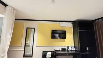 A hotel room with a bed, a desk with a chair, a flat-screen TV mounted on the wall, a mirror, and an air conditioning unit.