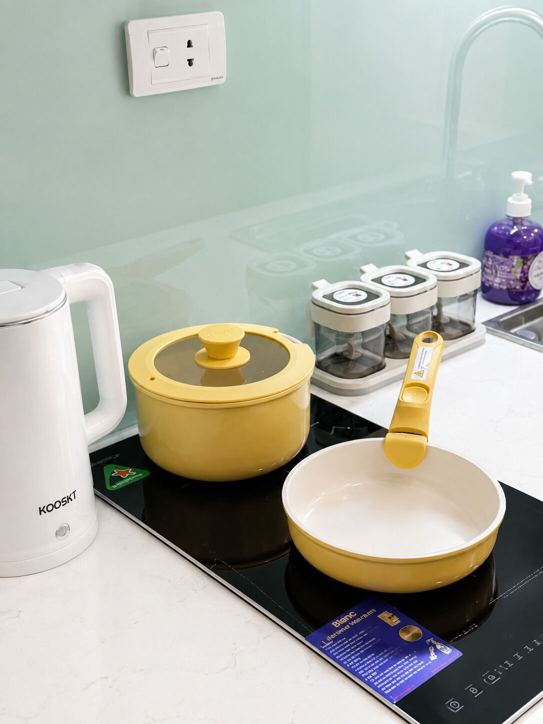 A kitchen with a white kettle, a yellow pot, and a pan on an induction cooktop. There are three spice containers and a bottle of dish soap on the counter.