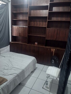 A bedroom with a bed, a white chair, a small table, and a large wooden shelving unit.
