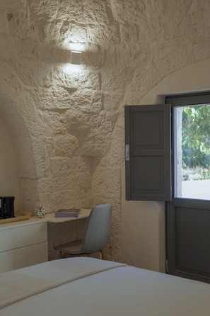 A room with a stone wall, a desk with a chair, a bed, and a window with a view of greenery. - Masseria Pastinaca Relais & Spa (Cisternino)
