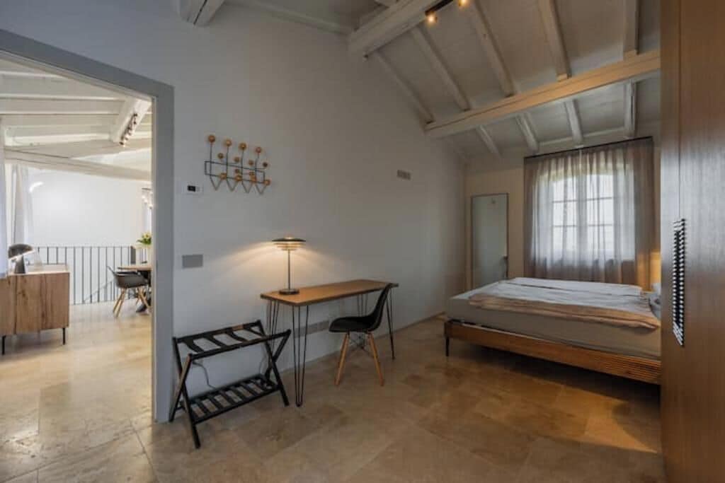 A bedroom with a bed, a desk, and a chair. There is a window with curtains and a door in the background.