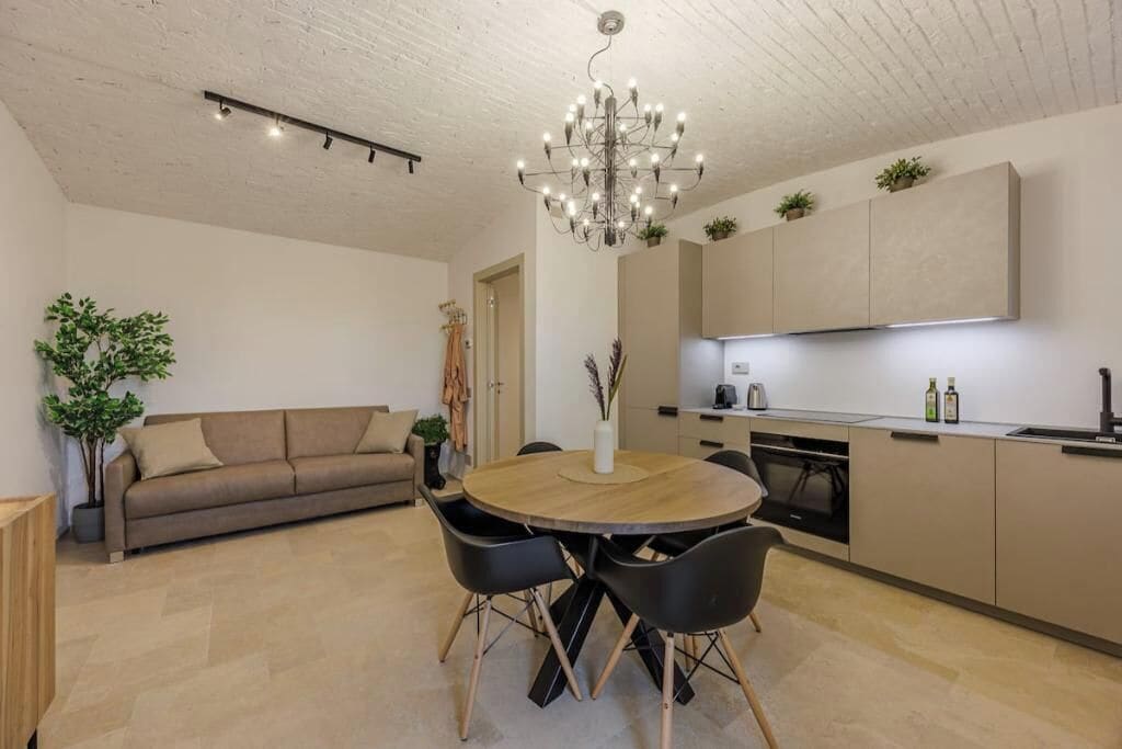 A modern kitchen with built-in appliances, a dining area with a round table and chairs, and a living room with a sofa and a plant.