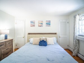 A bedroom with a bed, a dresser, a nightstand, and a window with curtains. - Connecticut Beach Home (Old Lyme)