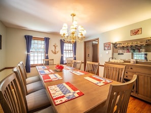 A dining room with a wooden table, chairs, a chandelier, and a kitchen area with a wine rack. - Connecticut Beach Home (Old Lyme)