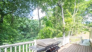 A barbecue grill on a wooden deck with a white railing, surrounded by lush greenery. - Connecticut Beach Home (Old Lyme)