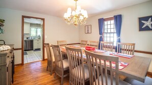 A dining room with a wooden table and chairs, a chandelier, and a view of the kitchen through an open doorway. - Connecticut Beach Home (Old Lyme)