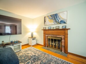 A living room with a fireplace, a flat-screen TV mounted on the wall, a lamp, and a patterned rug. - Connecticut Beach Home (Old Lyme)