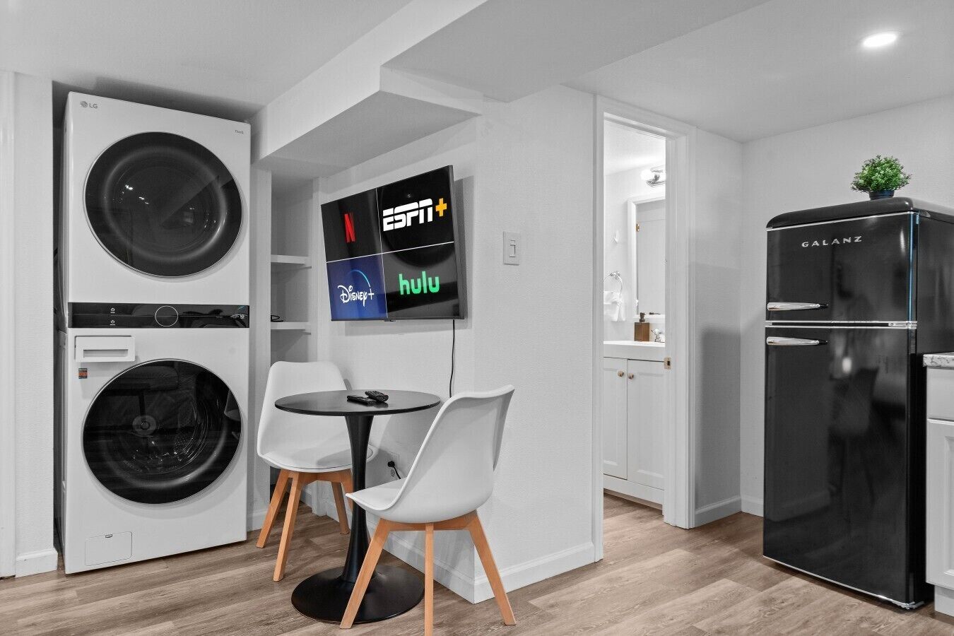 A compact kitchen with a washing machine, a black refrigerator, a dining table with chairs, and a wall-mounted TV displaying streaming services.