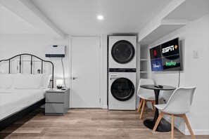 Ein Hotelzimmer mit einem Bett, einem Schreibtisch mit Lampe, einer Waschmaschine, einem Fernseher, der ESPN+ und Hulu zeigt, und einem kleinen Tisch mit Stühlen.