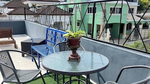 Terraza en la azotea con una mesa de vidrio redonda, dos sillas, una planta en maceta y vistas a edificios verdes y una montaña.