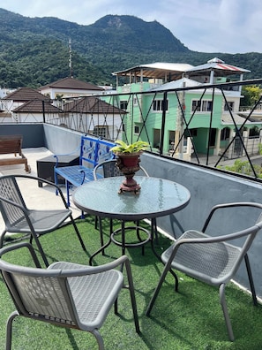 Eine Dachterrasse mit rundem Glastisch, zwei Stühlen, einem Blumentopf und Blick auf grüne Gebäude und einen Berg.