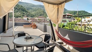 Un balcón con una mesa, sillas, una hamaca y vistas a una ladera verde con edificios.
