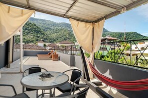 Ein Balkon mit Tisch, Stühlen, einer Hängematte und Blick auf eine grüne Hügelkuppe mit Gebäuden.