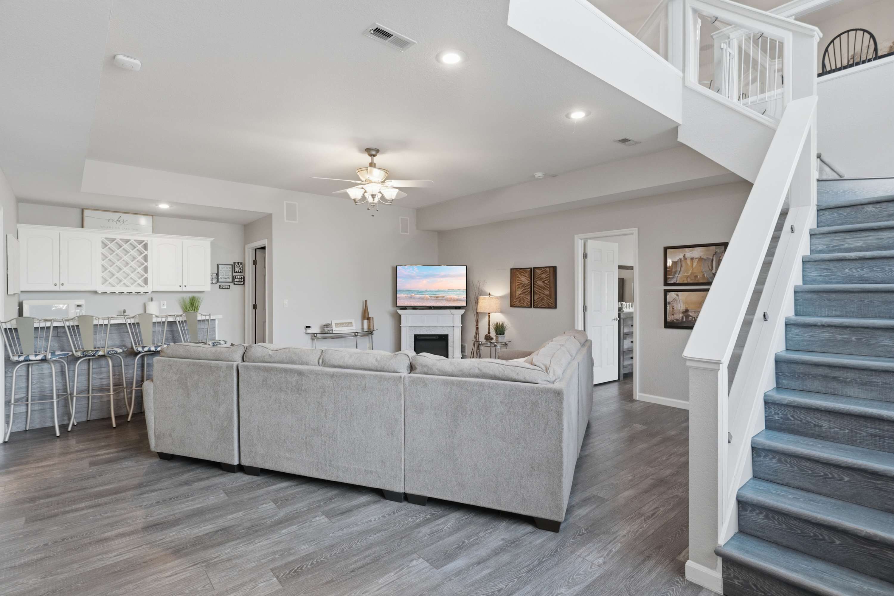 A spacious living area with a large grey sectional sofa, a dining area with a white kitchen in the background, and a staircase with grey steps.