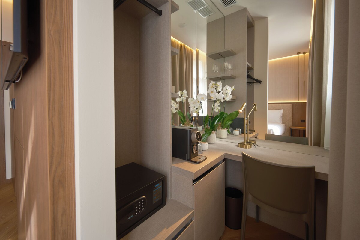 Una habitación de hotel moderna con una puerta de madera, un minibar, un escritorio con lámpara, una silla y un espejo grande que refleja la habitación.