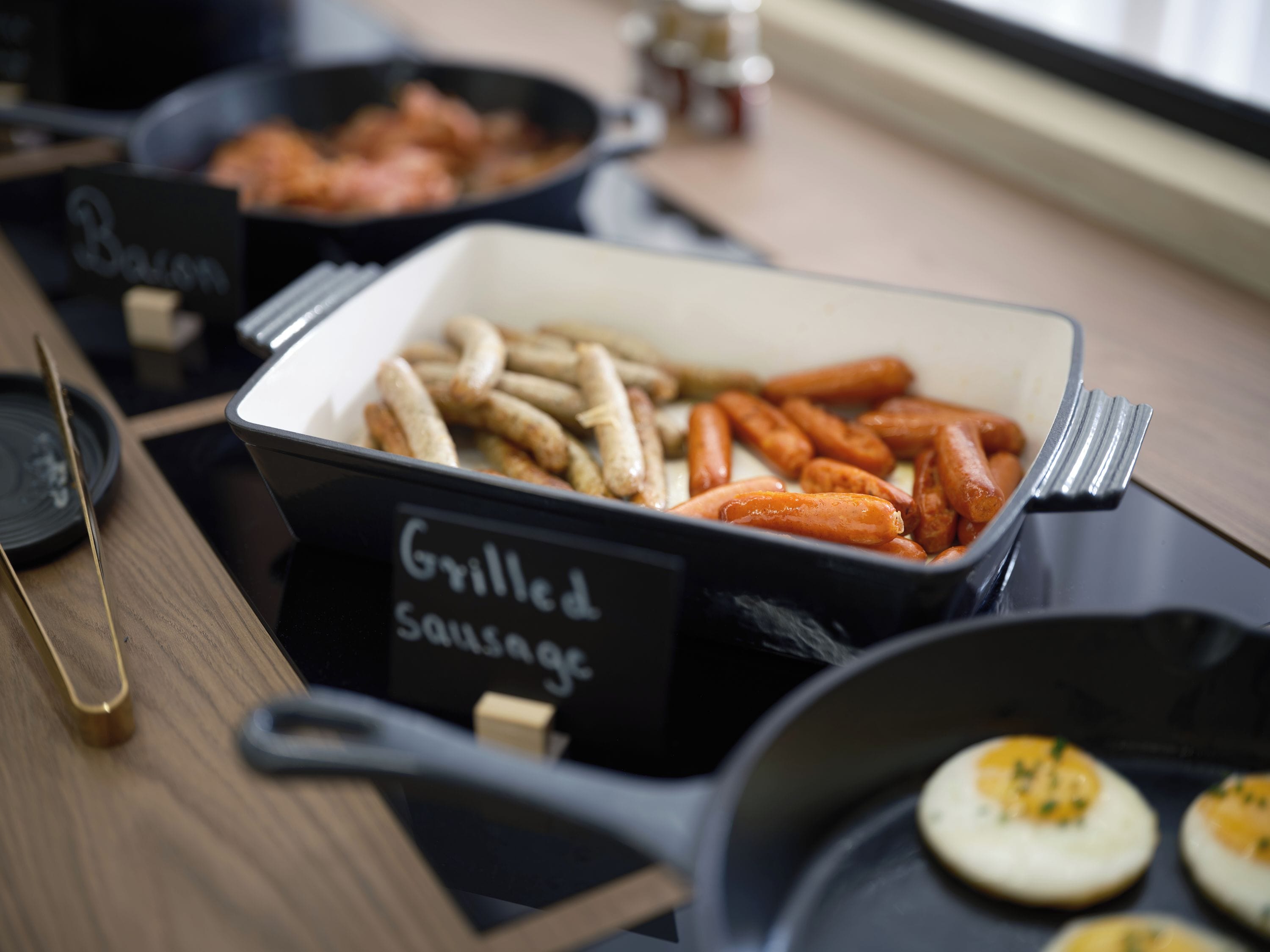 A buffet table with labeled containers of grilled sausages and bacon.