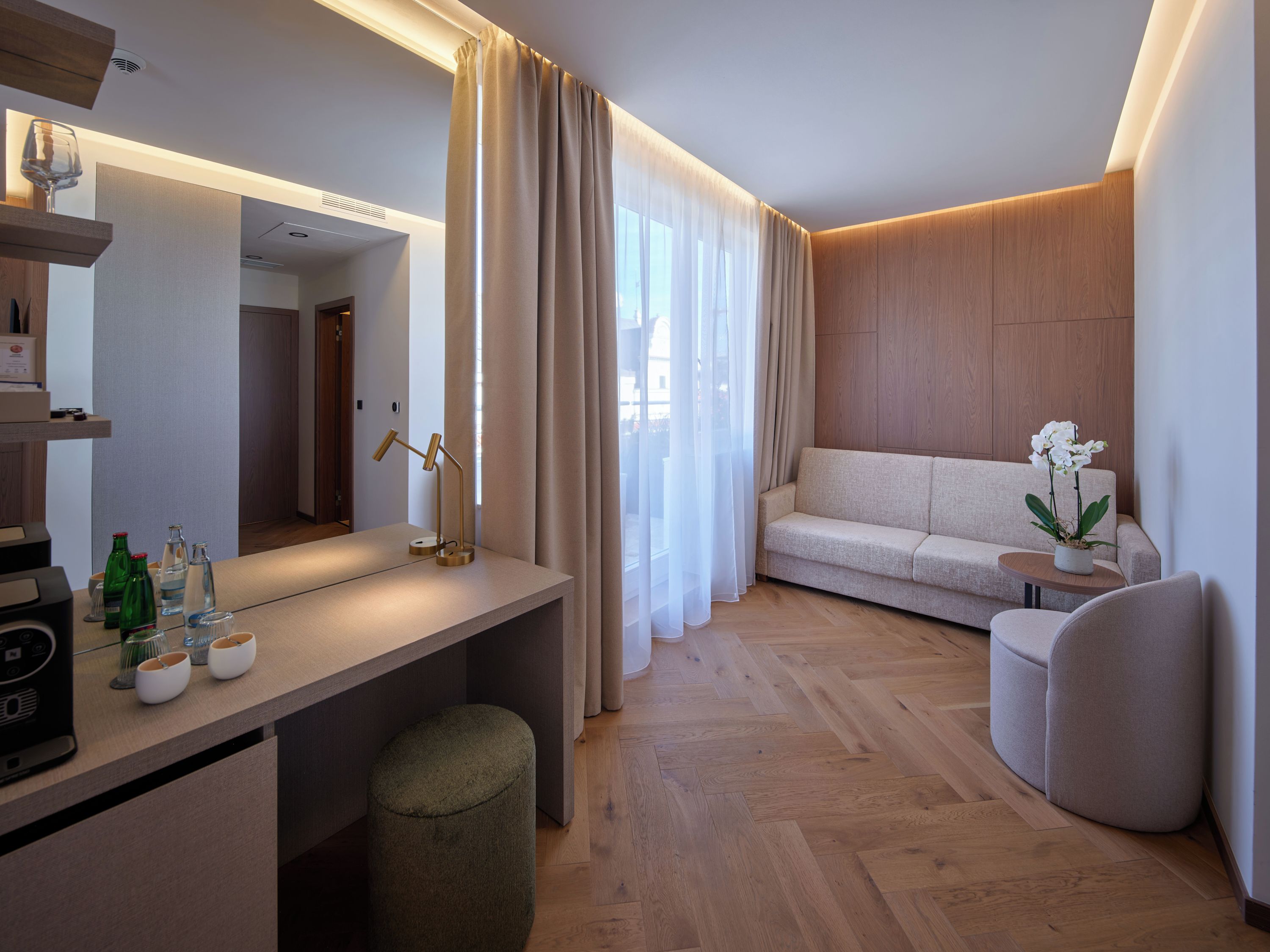 Een moderne hotelkamer met een houten vloer, een bank, een kleine tafel met een plant, een bureau met een lamp en een grote spiegel.