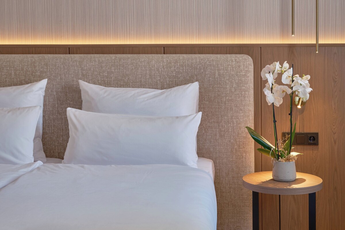 Habitación de hotel con cama, almohadillas blancas, una mesita redonda con orquídea en maceta y una pared de madera con líneas verticales.