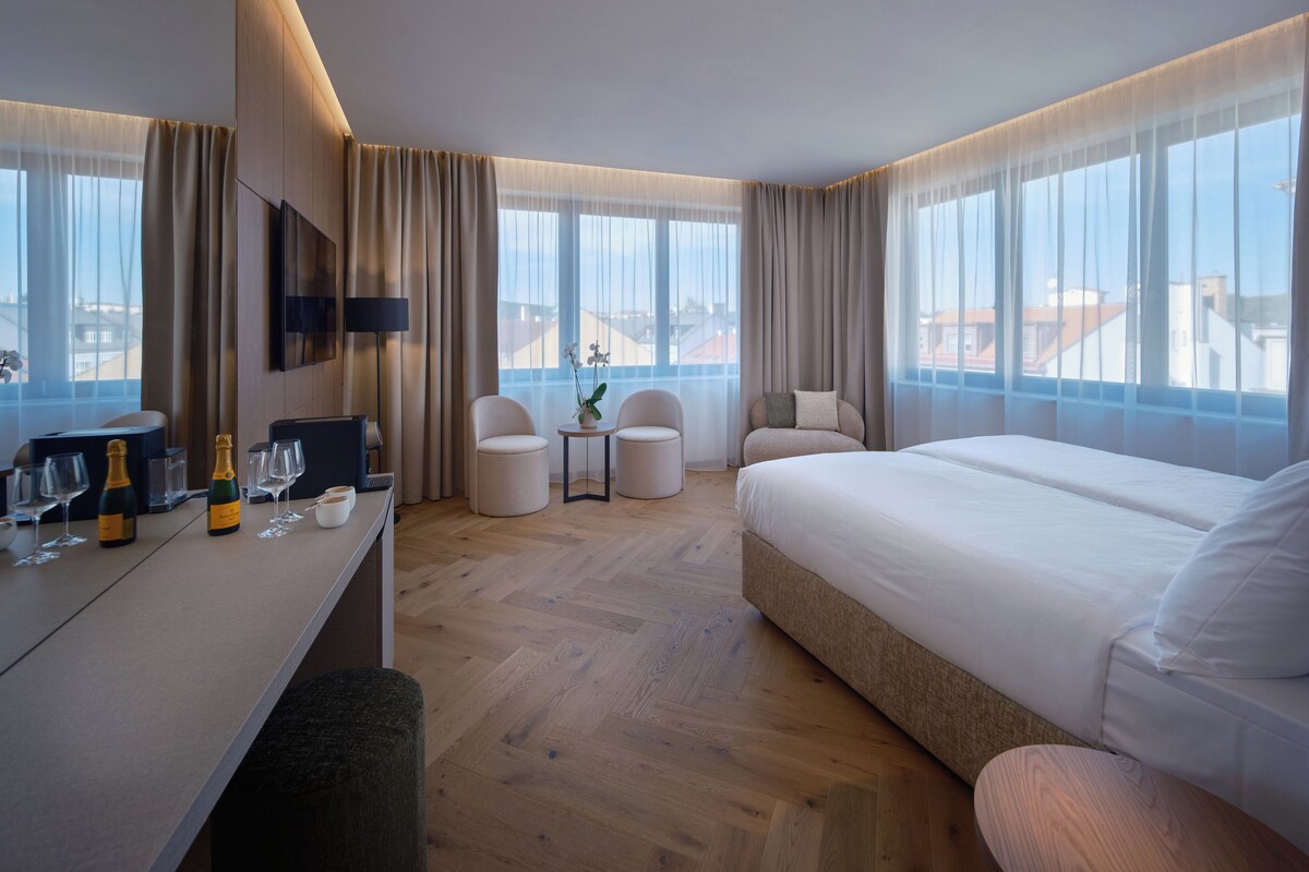 Una habitación de hotel con una cama, dos sillones, una mesita con un florero y un escritorio con vasos y una botella.
