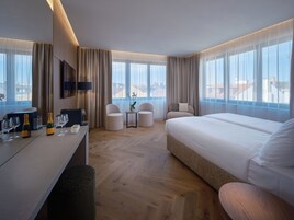 Quarto de hotel com uma cama, duas poltronas, uma mesinha com um vaso e uma escrivaninha com copos e uma garrafa.