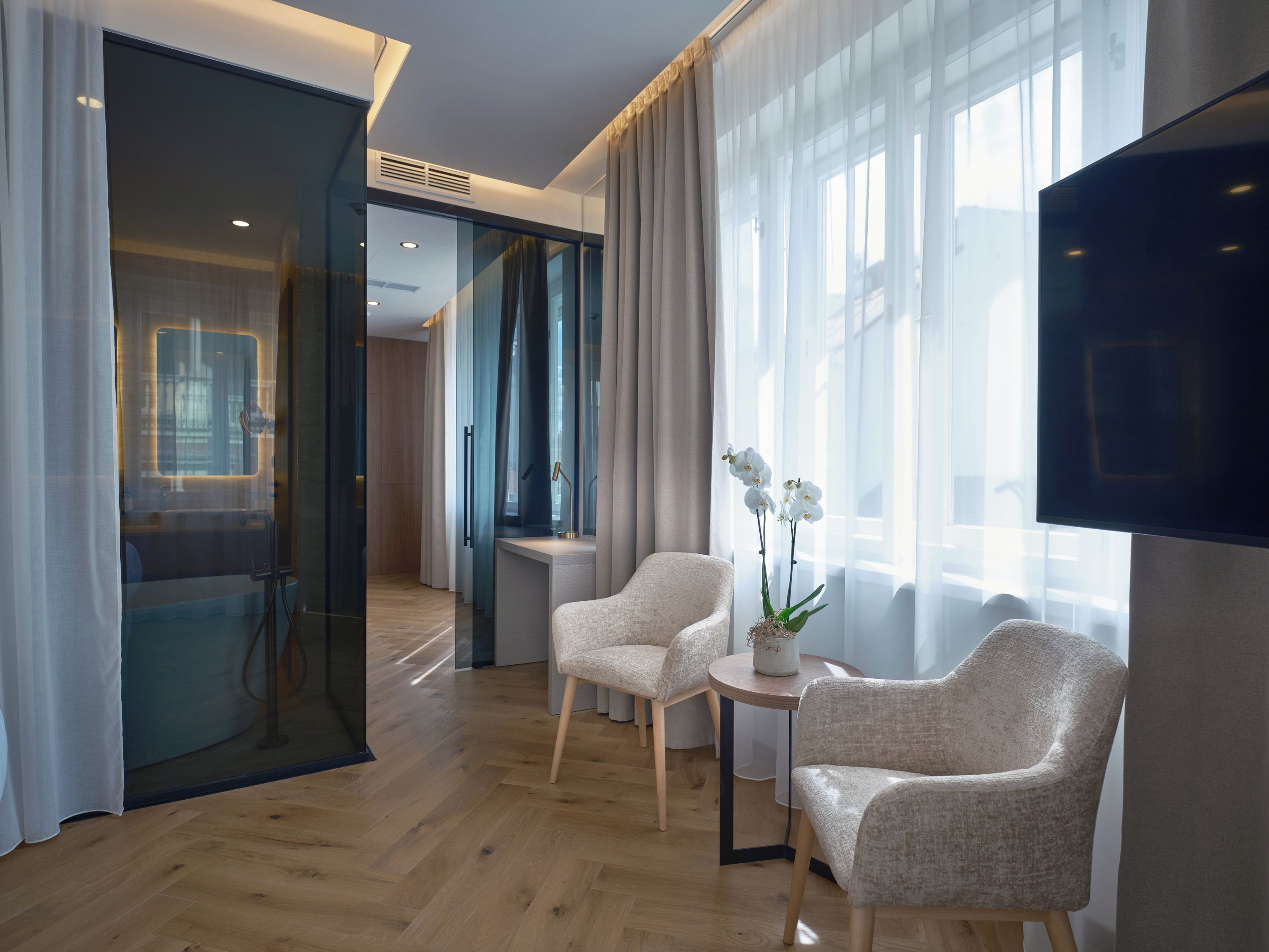 Quarto de hotel moderno com piso de madeira, uma janela grande com cortinas leves, televisão de tela plana e duas poltronas estofadas.