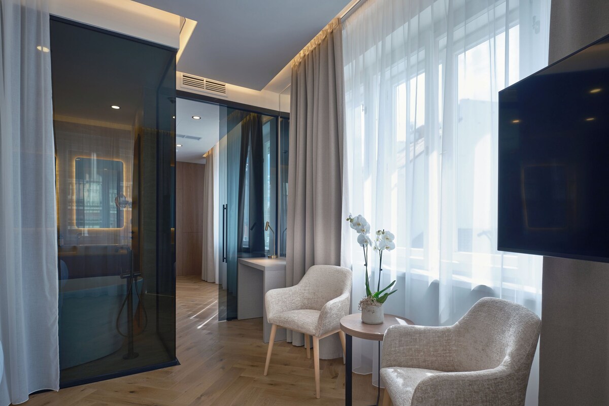 Una habitación de hotel moderna con piso de madera, un ventanal con cortinas translúcidas, un televisor de pantalla plana y dos sillones tapizados.