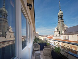Un balcón con sillones de mimbre y una pequeña mesa, con vistas a un paisaje urbano que incluye edificios históricos y una torre destacada.