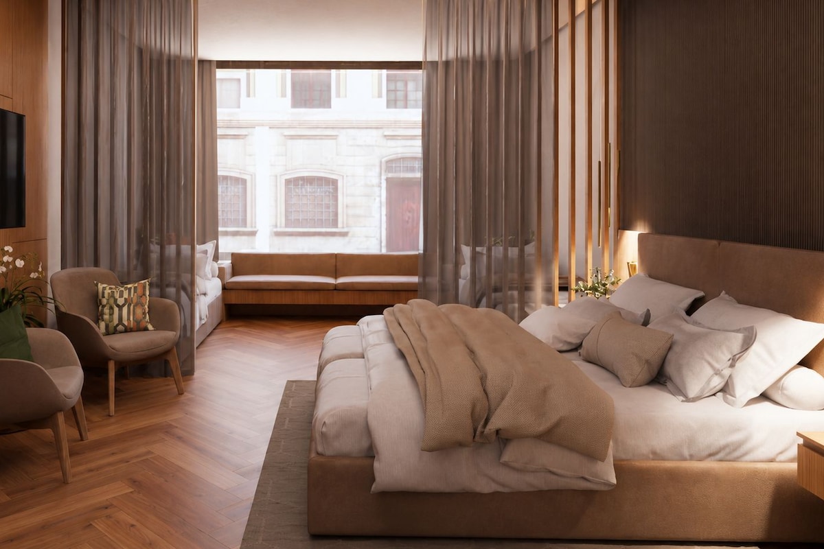 A modern bedroom with a large bed, a seating area with a chair and sofa, and a view of a building through a window.