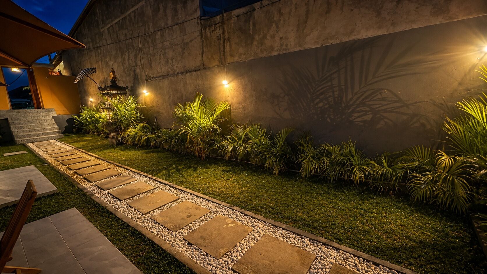 A well-lit garden pathway with stepping stones, surrounded by lush greenery and a decorative wall.