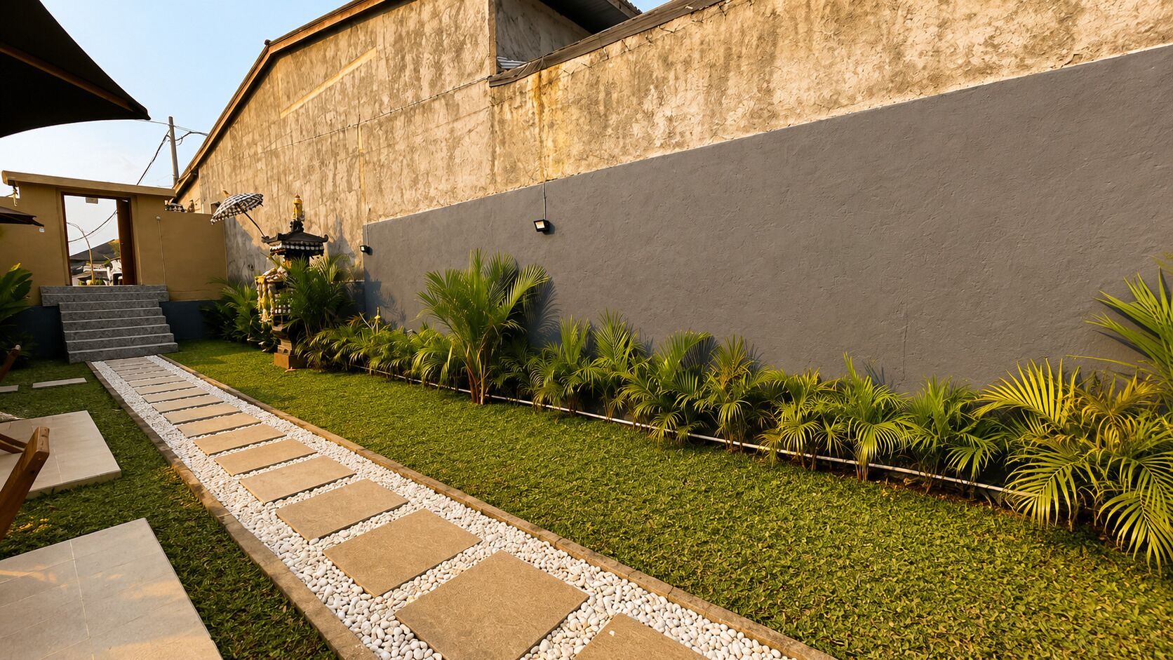 A pathway leading to a building with a modern design, a small garden area with palm trees, and a clear sky.