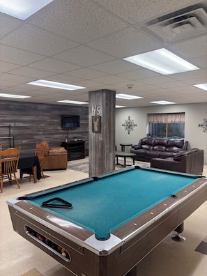Une table de billard dans une pièce dont un mur est en bois, avec un canapé en cuir et un téléviseur fixé au mur.