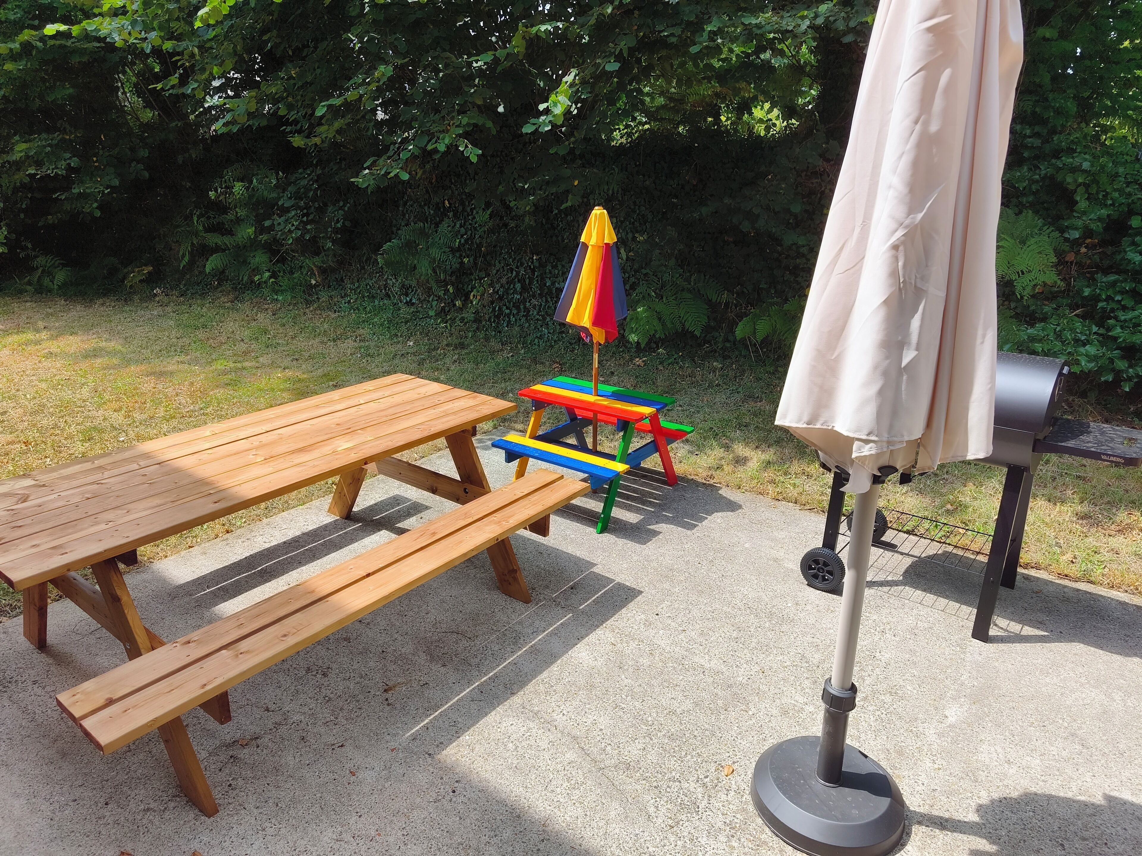 A picnic setup with a wooden table and benches, a colorful play table, a grill, and an umbrella.