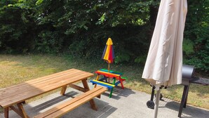 A picnic setup with a wooden table and benches, a colorful play table, a grill, and an umbrella.