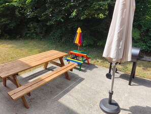 Montaje para picnic con una mesa y bancos de madera, una mesa de juego colorida, una parrilla y una sombrilla.